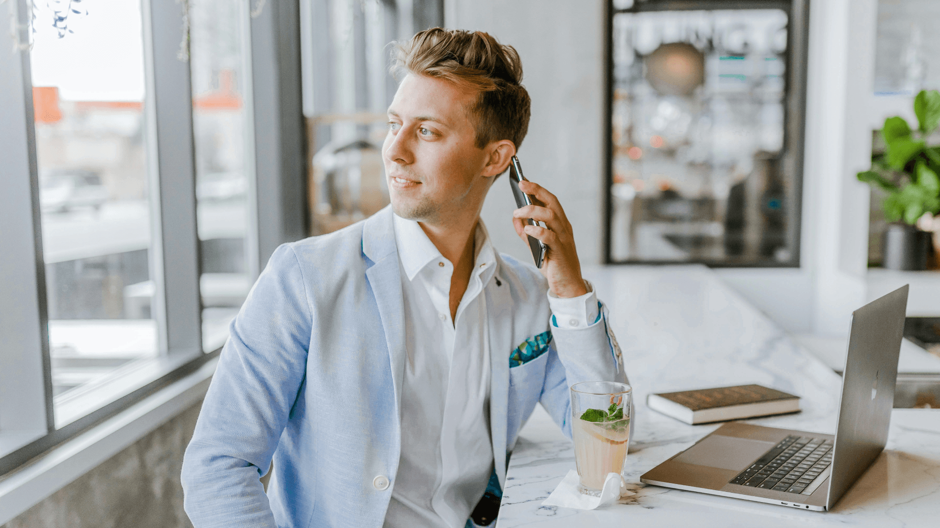 Man in a light blue blazer working on a laptop while speaking on his cell phone.